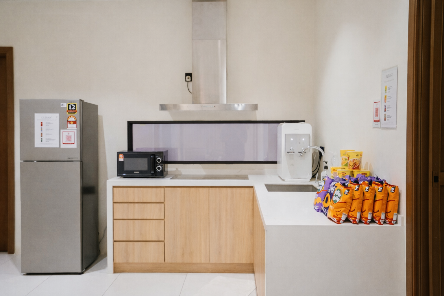 A photo of a kitchen with a fridge, microwave, kitchen hood exhaust fan, water dispenser and a wash basin. Snacks are placed on the countertop.